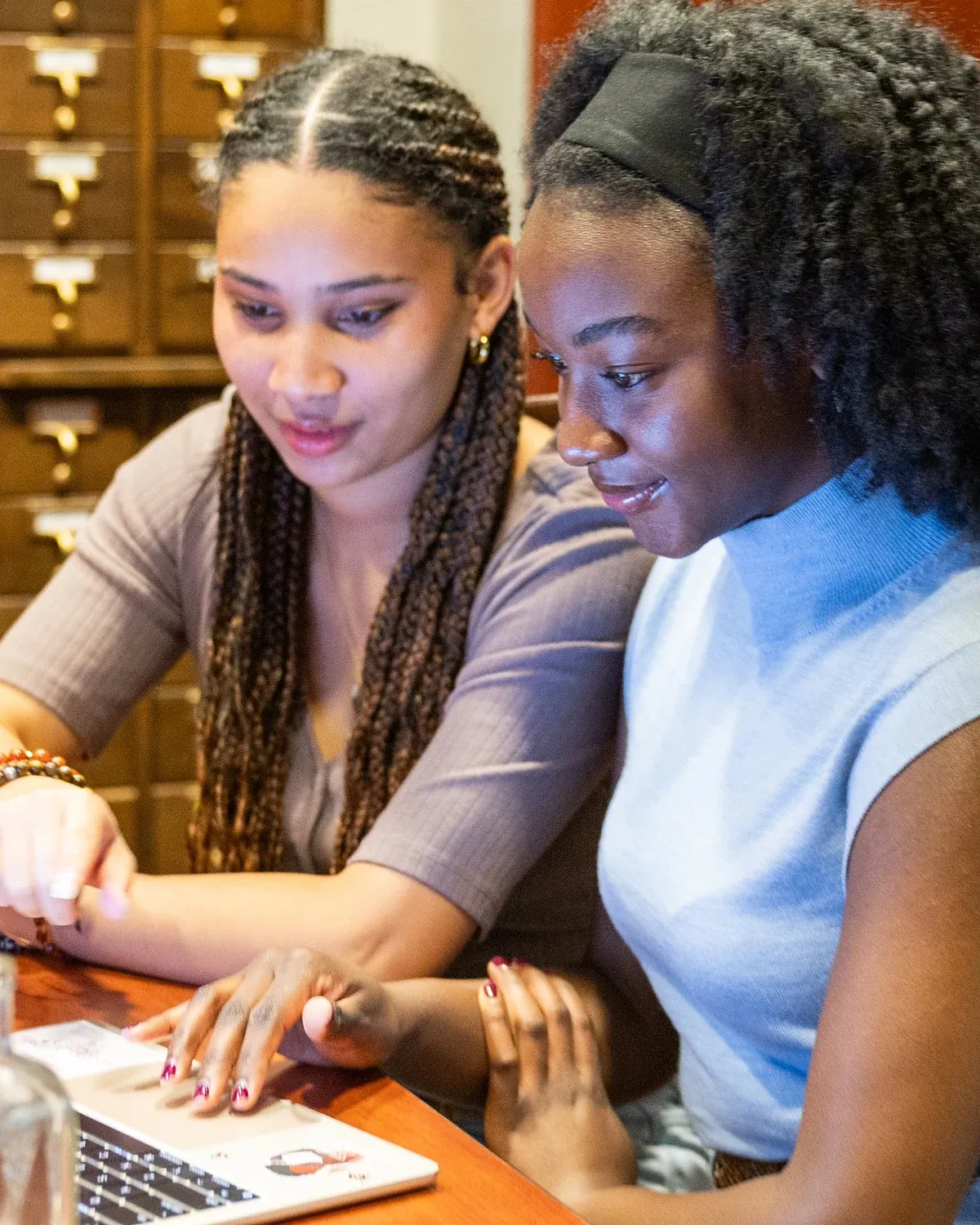 Two students from the BWDP team working together at a laptop in the library, with wooden card-catalog drawers behind them and an antique glass bottle on the table